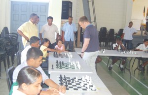 Chess Grandmaster Rainer Buhmann (centre) competes with one of his  challengers during a display of facing multiple opponents yesterday at QC,  while Prime Minister Samuel Hinds (standing left) appreciates the moment. 