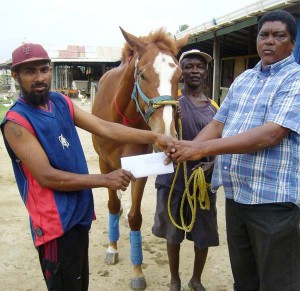 Boyo Mohabir (left) of the organising committee receives the cheque from Glendon Sooklall of the Shariff Racing stable. Groom of the stable William LaRose holds top horse Spit Fire in the background.