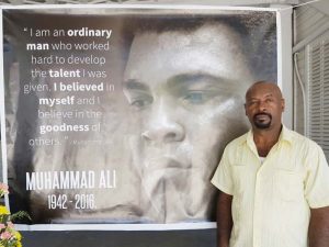 Max Massiah stands beside a poster of Muhammad Ali at the Ministry of Culture, Youth and Sports on Main Street where a book of condolence has been opened.